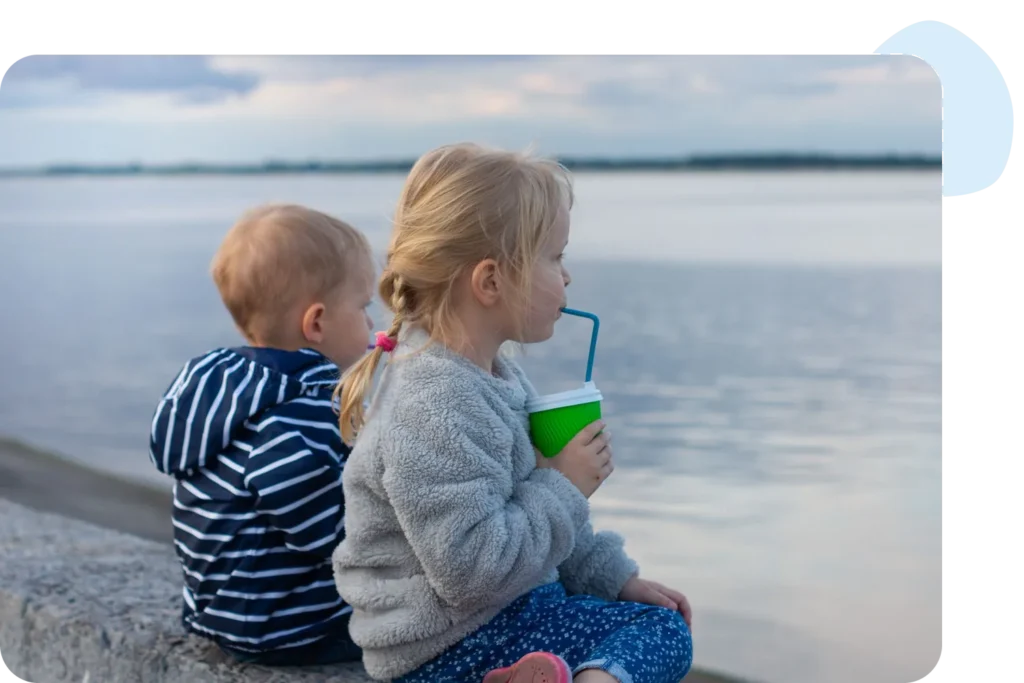 Two children sit by a lake, one drinking from a cup, enjoying a peaceful day outdoors.