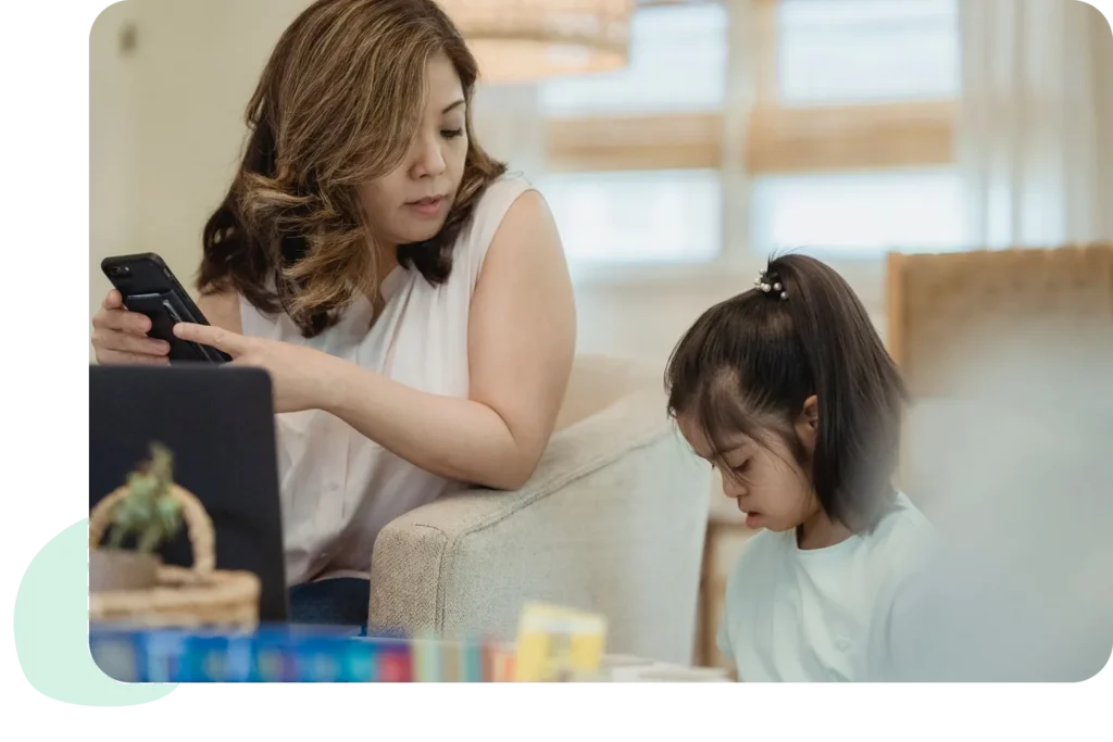 A mother using a smartphone while her daughter reads nearby, creating a warm family moment.
