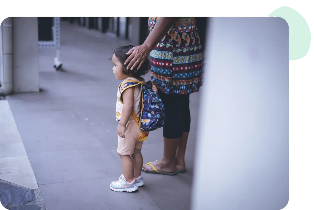 A young child with a backpack stands beside a caregiver's protective touch outdoors.