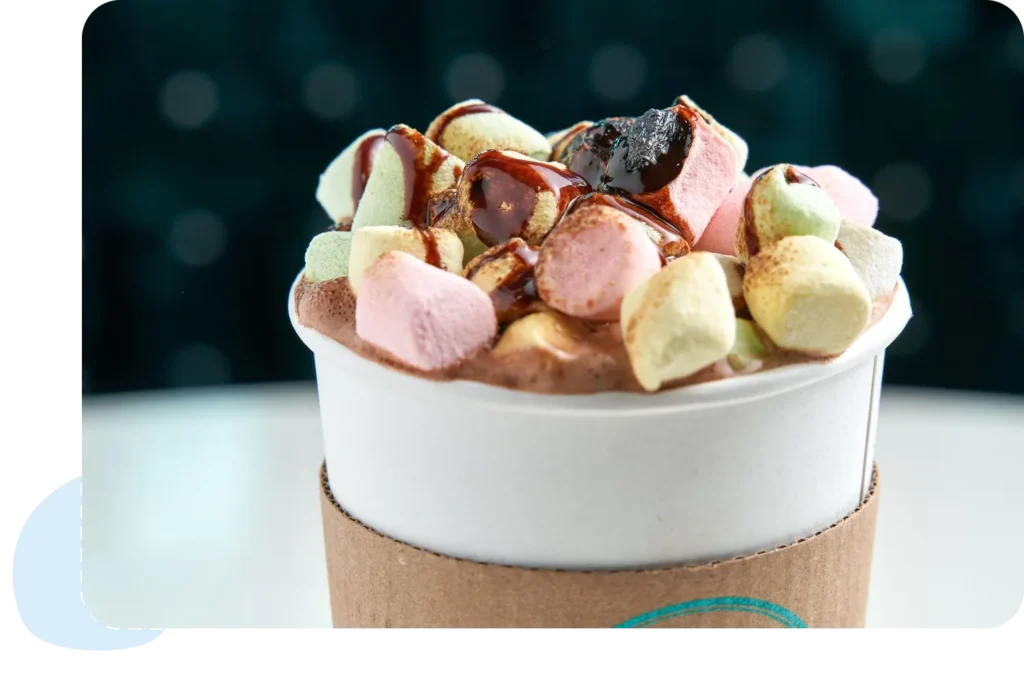 Close-up of hot chocolate topped with colorful marshmallows and chocolate syrup in a to-go cup.
